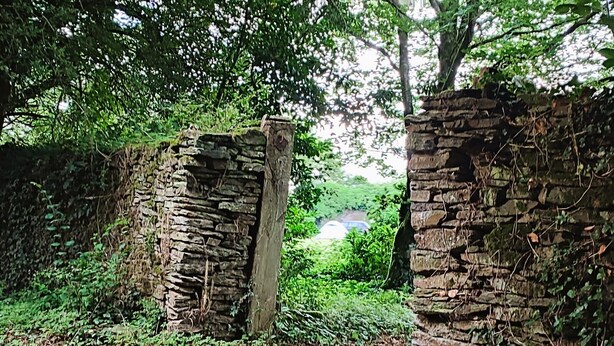 Una rústica puerta de piedra da paso a una zona boscosa con un sendero y una vista distante de un coche azul.