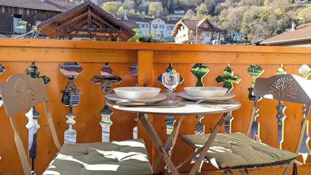 Un balcon en bois avec une table dressée pour le dîner, offrant une vue sur un paysage montagneux parsemé de maisons.