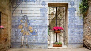 Eine mit Fliesen verkleidete Wand mit einem Wandgemälde, das eine Figur mit Stab zeigt, ein Tor mit einem dekorativen Schlüssel und ein Blumenbeet.