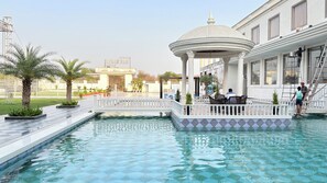 A pool area with a gazebo, palm trees, and a building with large windows. - ORCHHA RIVER STONE RESORT (Nivari)
