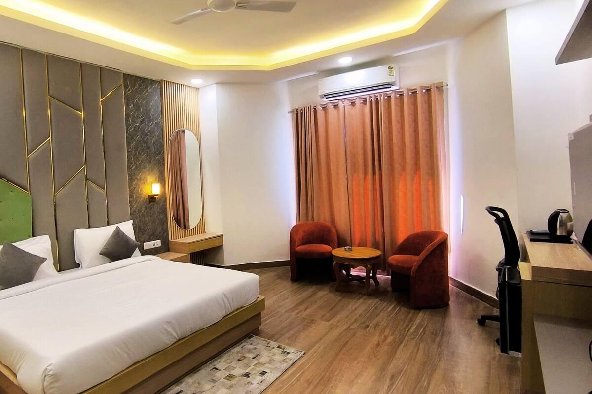Une chambre d’hôtel moderne avec un grand lit, deux fauteuils rouges, une petite table ronde et un bureau avec un ordinateur.
