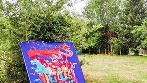 Et farverigt banner med tekst og illustrationer, placeret på græs i et udendørs miljø med træer i baggrunden.