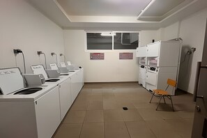 A laundry room with multiple washing machines and dryers, a window, and a chair.