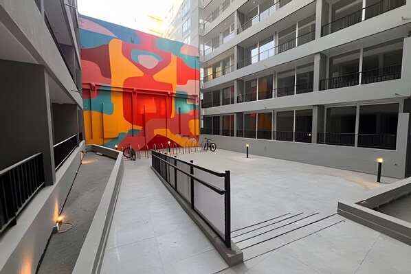 A modern building with a colorful mural on the side, a covered outdoor area with a bike rack, and a spacious courtyard with a railing.
