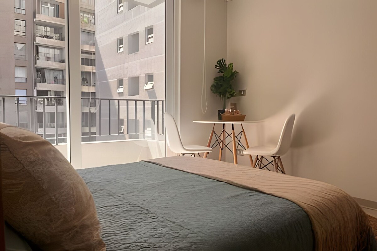 A bedroom with a bed, a small table with chairs, a potted plant, and a view of buildings through large windows.