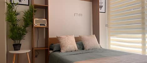 Une chambre à coucher avec un lit, une table de chevet, une plante en pot, une radio et une fenêtre avec des stores.