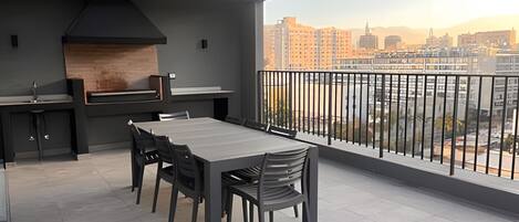 A modern outdoor dining area with a large table and chairs, a built-in grill, and a view of a cityscape.