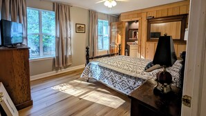 Ein Schlafzimmer mit Holzboden, einem Bett mit gemustertem Überwurf, einem Deckenventilator, einem Fernseher auf einer hölzernen Kommode und Blick ins Freie durch große Fenster.
