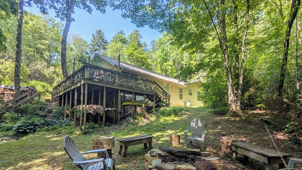 Eine rustikale Holzhütte mit Veranda, umgeben von einem Wald. Adirondack-Stühle und eine Feuerstelle sind vorhanden.