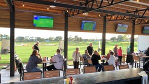 Ein Außenbereich mit Tischen und Stühlen, Blick auf einen Golfplatz und mehrere Bildschirme, die Sportübertragungen zeigen.