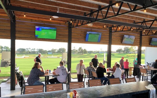 An outdoor seating area with tables and chairs, a view of a golf course, and multiple screens displaying sports content.