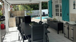 Et overdækket spiseområde med bord og stole, grill og udsigt til en swimmingpool.