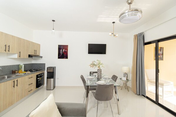 A modern kitchen with wooden cabinets, a built-in oven, a coffee machine, and a dining area with a glass table and chairs.