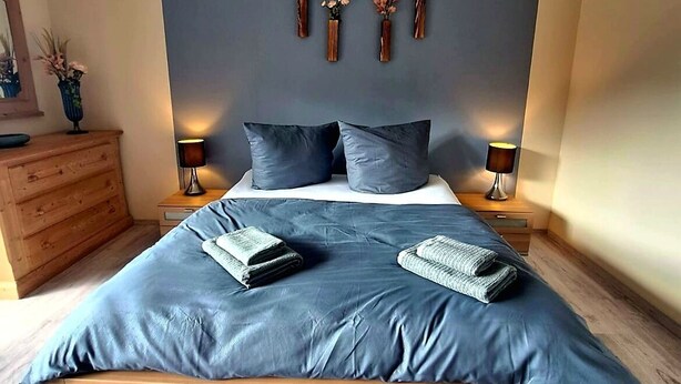 A bedroom with a large bed, two bedside tables, a wooden dresser, and a bench. - Charming, family-friendly vacation apartment in the heart of the Volcanic Eifel (Niederstadtfeld)