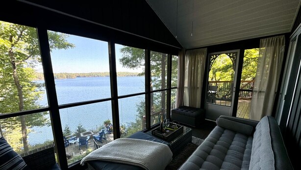 Una habitación con un ventanal grande con vistas a un lago, un sofá, una mesita con una botella y un banco acolchado.