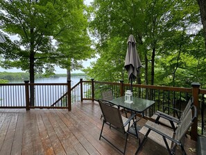 A wooden deck with a glass table and chairs, overlooking a lake surrounded by trees. - 3 BR Lakefront Beach Retreat; Hot Tub, Fire Pits! Pet-friendly! (South Frontenac)