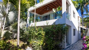 Een modern huis met twee verdiepingen en een balkon, omringd door tropische planten en een stenen pad.