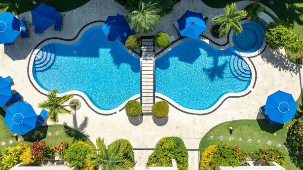 A large outdoor swimming pool with two separate sections connected by a bridge, surrounded by palm trees and blue umbrellas. - Pro Cab Paradise Condos Cabarete (Cabarete)