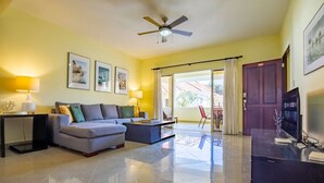 A modern living room with a sofa, coffee table, and TV. There is a ceiling fan, a door, and a window with curtains. - Pro Cab Paradise Condos Cabarete (Cabarete)