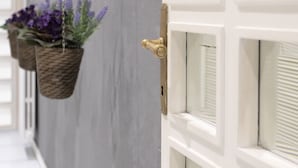 Un couloir avec une porte blanche, un panier suspendu contenant des fleurs violettes et un mur gris à la texture particulière.