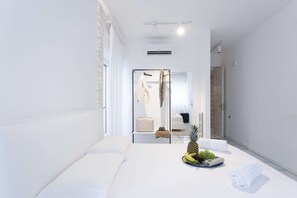A modern hotel room with a large bed, a small table with fruits and towels, and a view of the hallway and another room. - Bocà Home (Salerno)