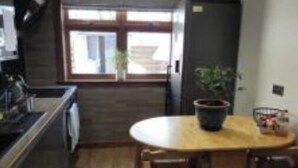 Une cuisine avec une table et des chaises en bois, un réfrigérateur et une fenêtre donnant sur un bâtiment à l’extérieur.