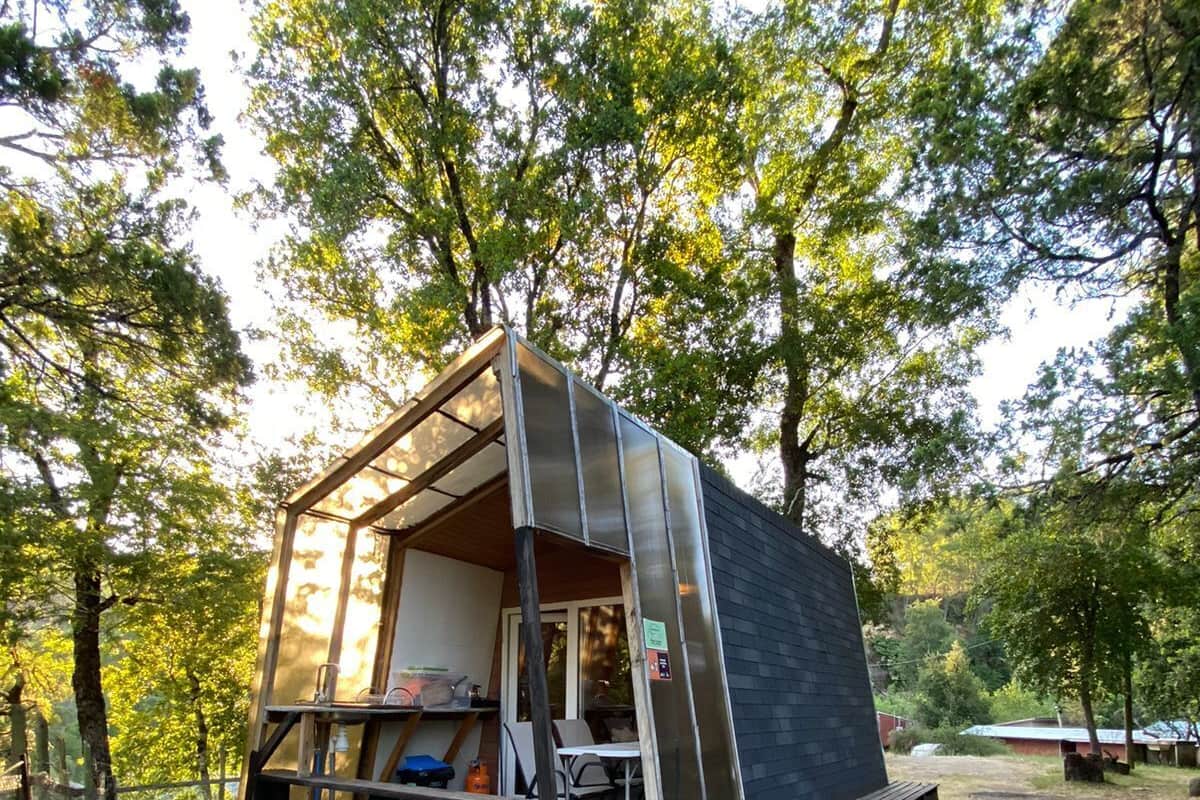 A small, modern cabin with a glass front, a wooden deck, and a metal grill.