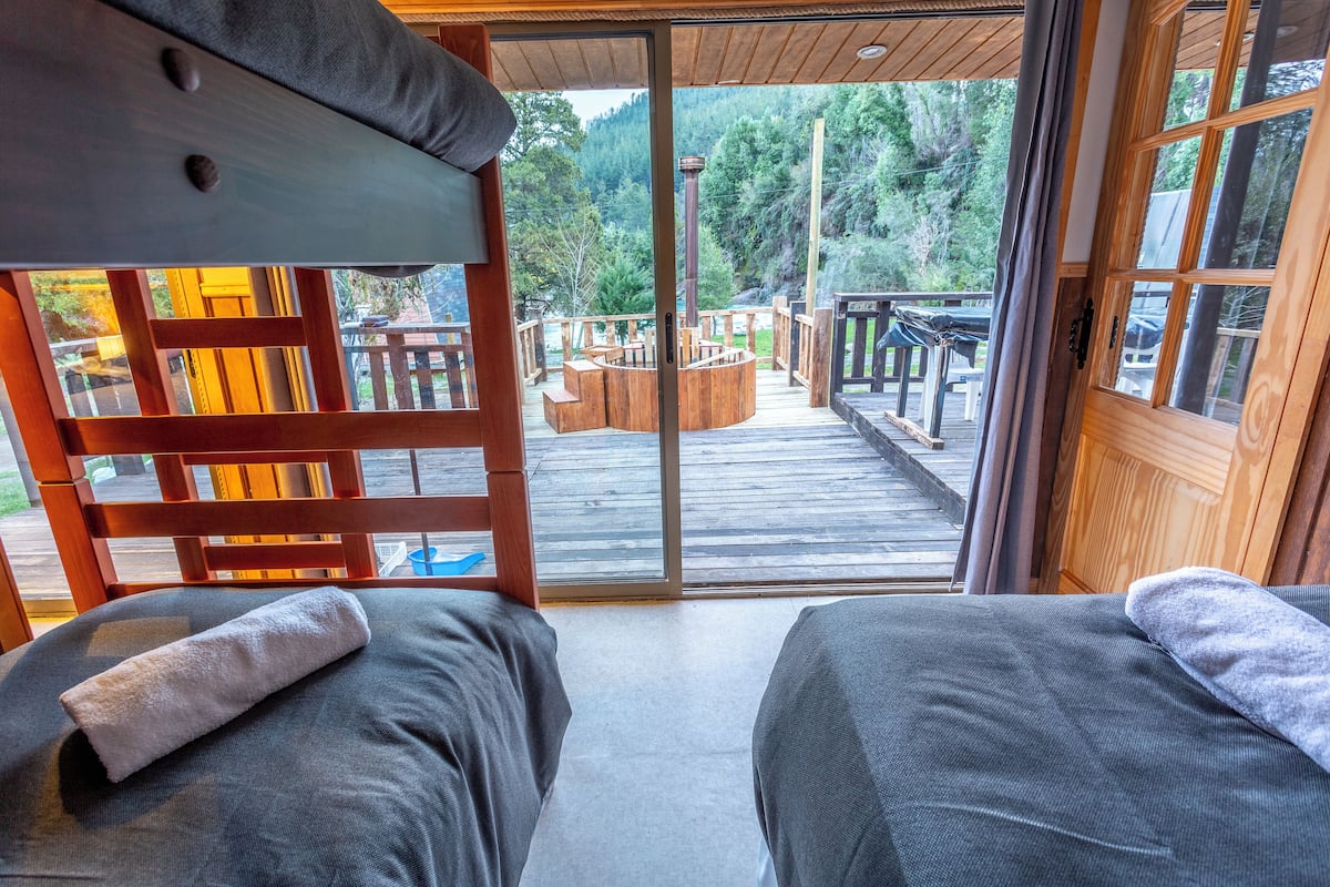 A room with two beds, a wooden deck outside, and a view of a forested area.