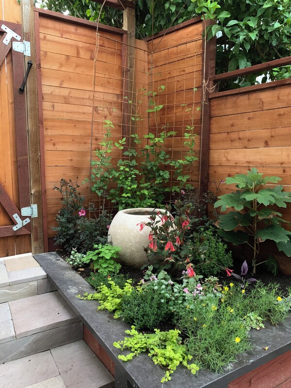 Un jardín de madera con una jardinera grande, escalones que llevan a ella, y diversas plantas y flores.