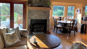 A cozy living room with a stone fireplace, wooden furniture, and large windows offering a view of the outdoors.