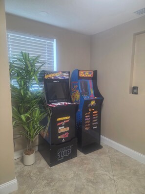 Two arcade machines labeled 'Asteroids' and 'Pac-Man' in a corner with a potted plant and a window with blinds.