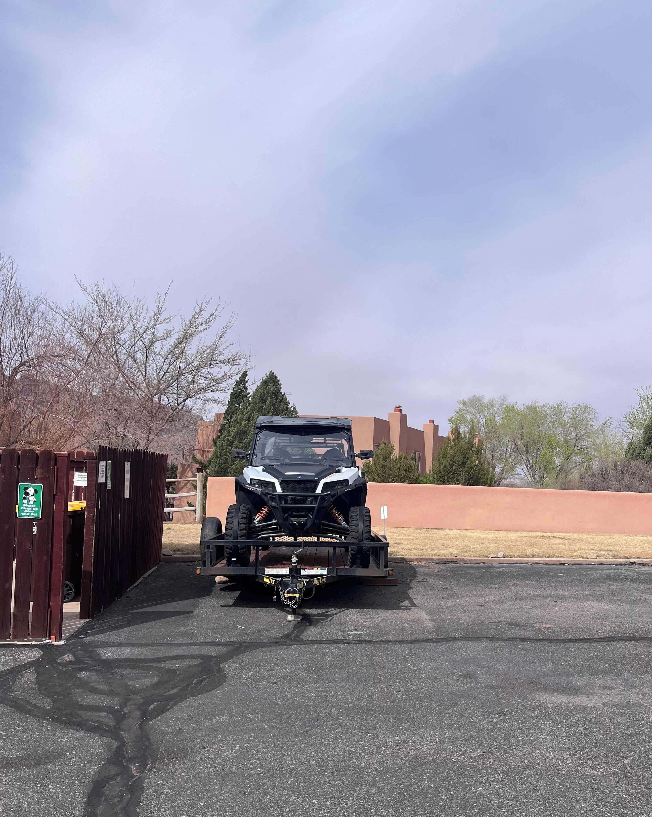 Voertuig op een aanhanger in een parkeerplaats, met een houten hek en een bord links.