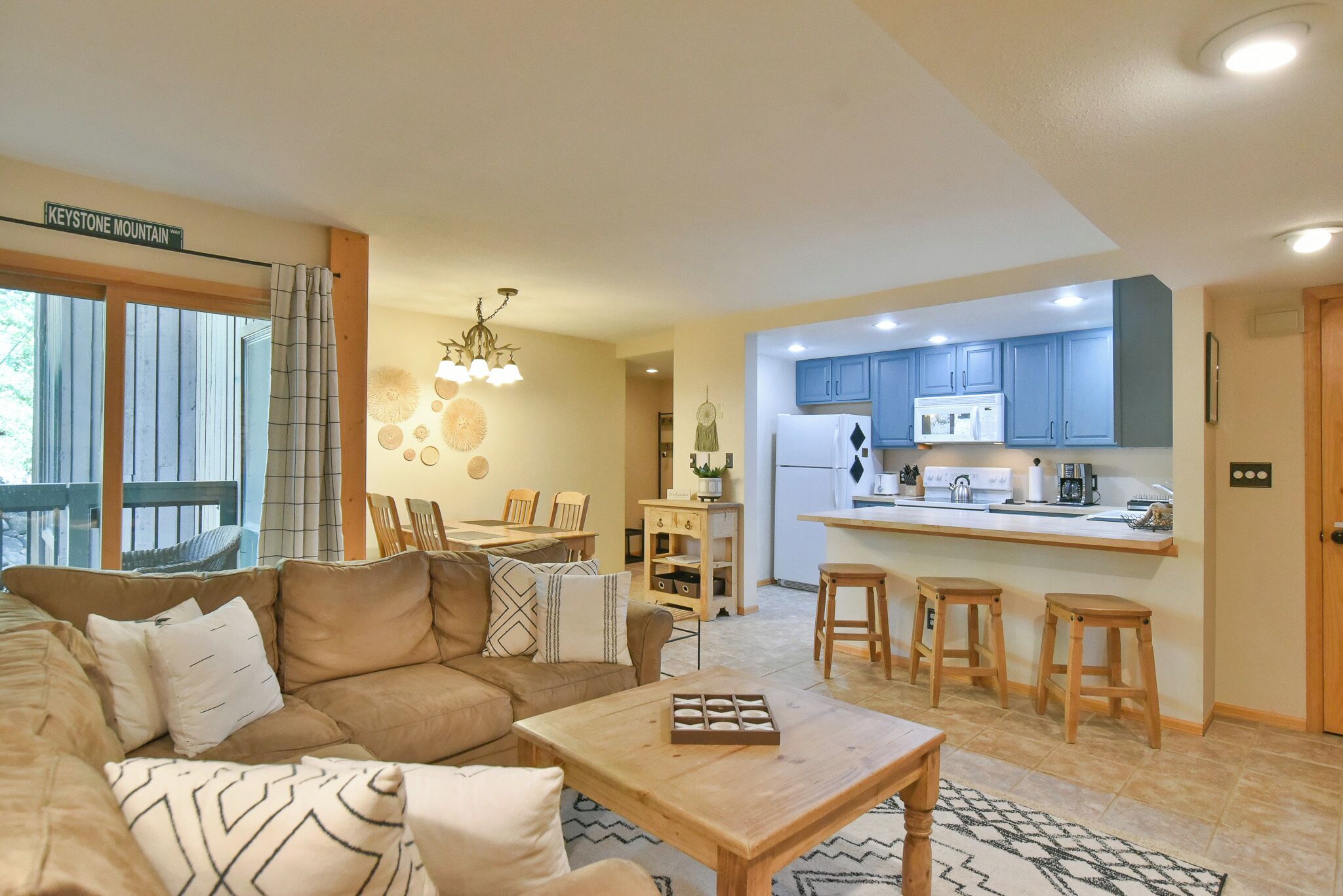 A cozy living room with a sofa, coffee table, and a dining area with chairs. A kitchen with blue cabinets and a refrigerator is visible.