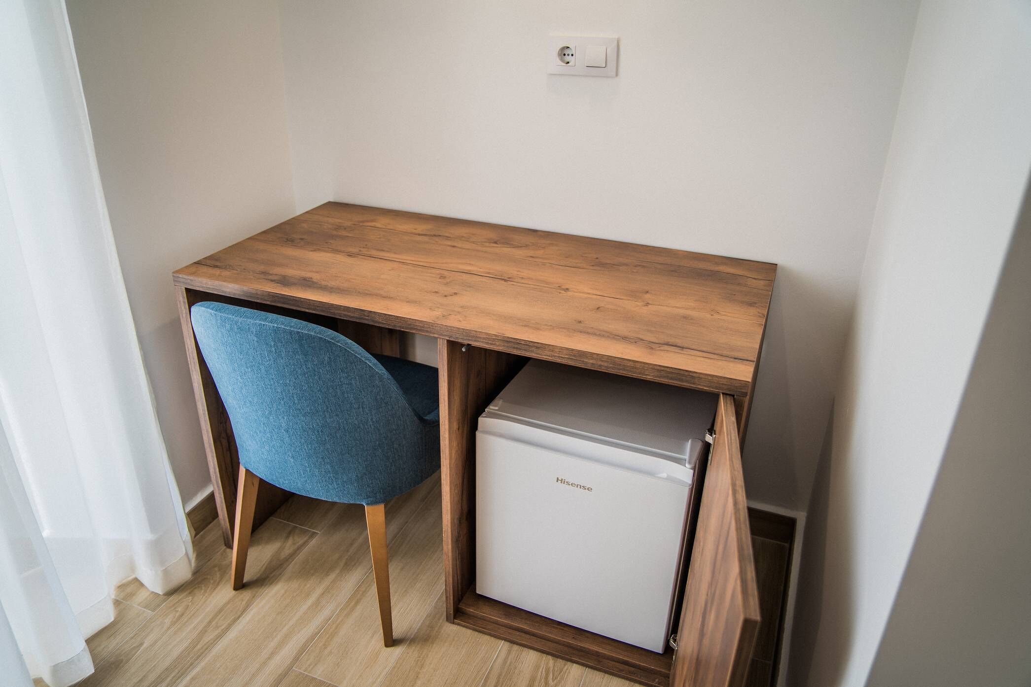 A compact room with a wooden desk, a blue chair, and a white mini-fridge.