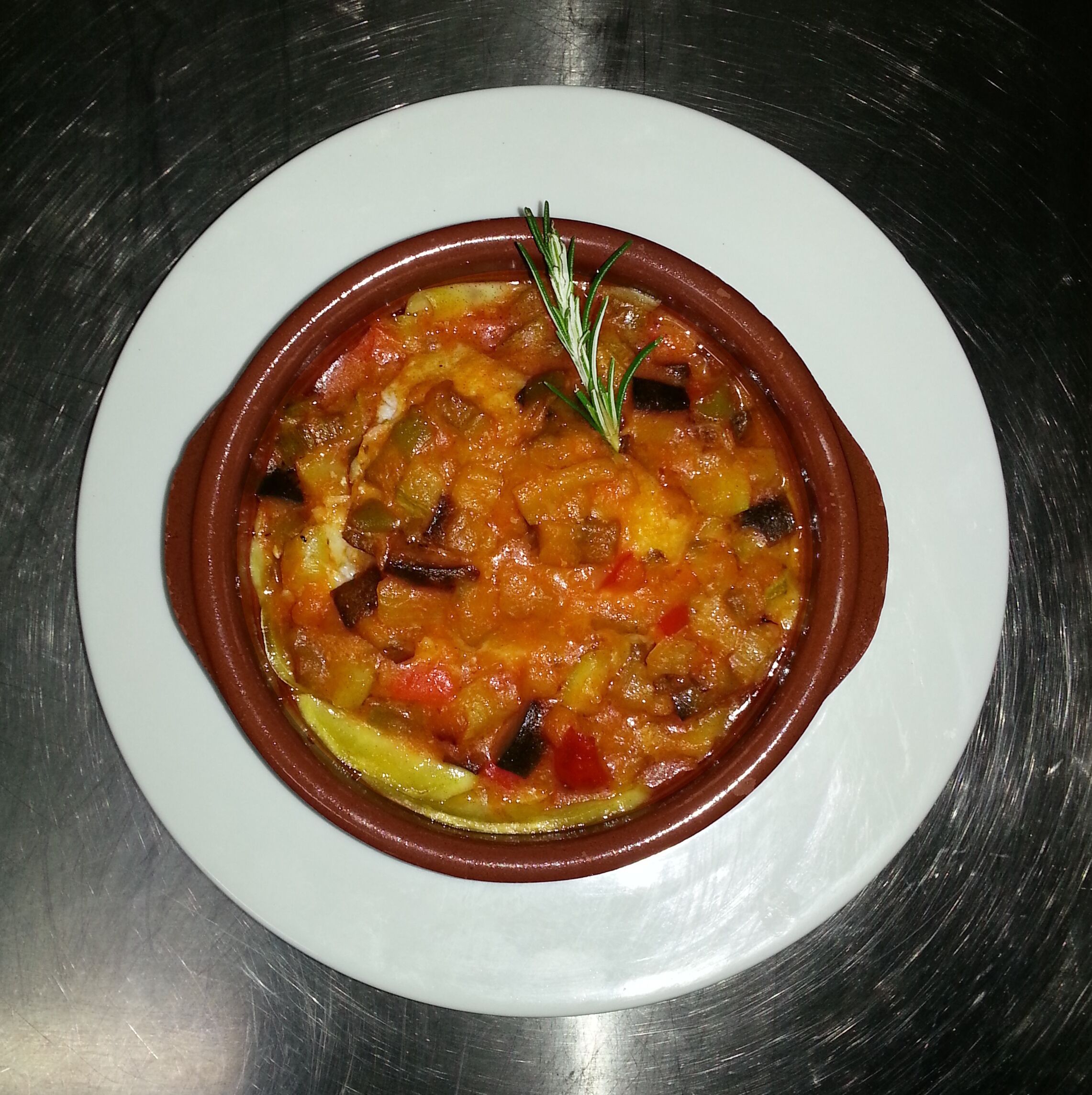 Un plato blanco con un cuenco de guiso, verduras y una ramita de romero.