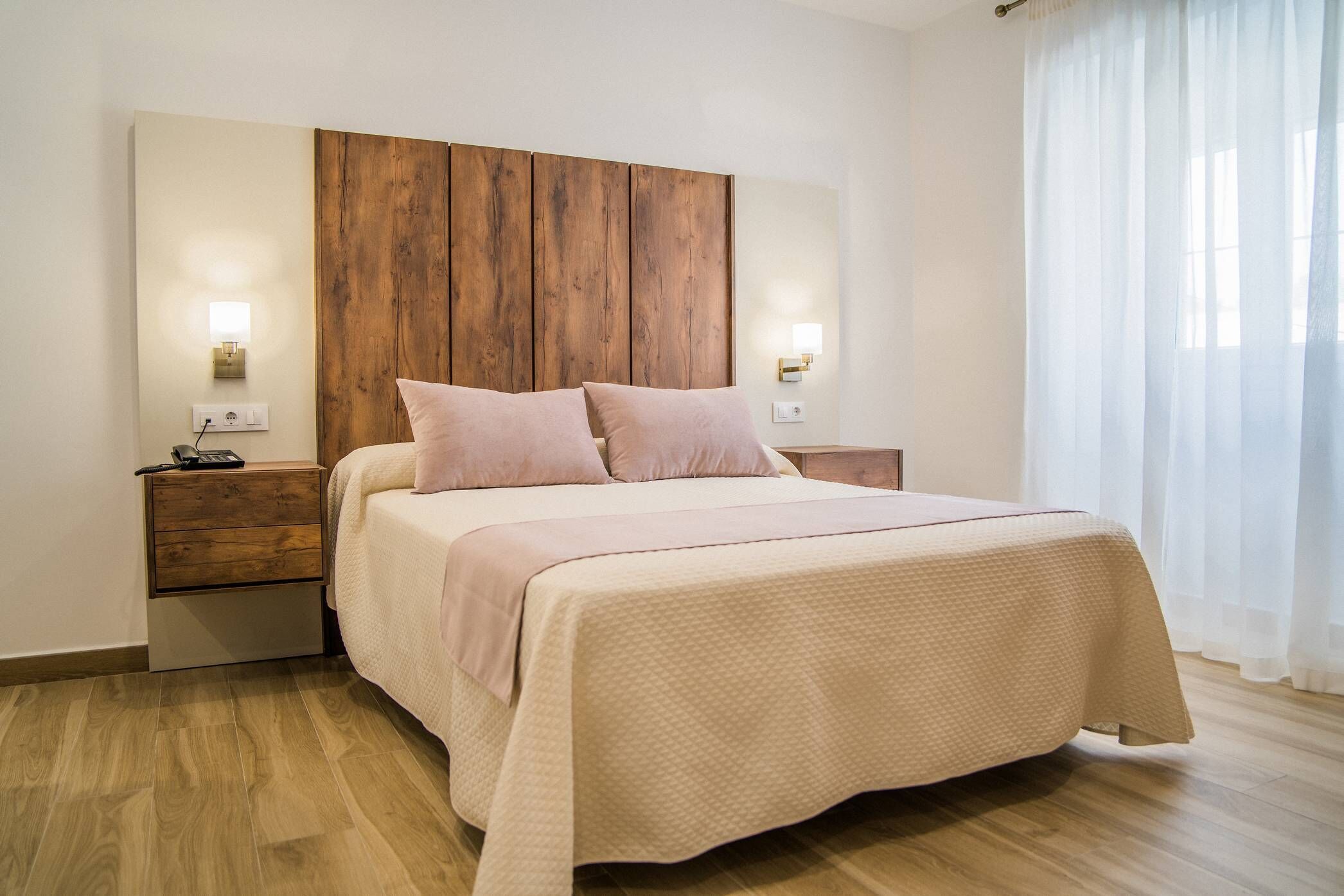 Un dormitorio con cabecera de madera, dos mesitas de noche con lámparas, una cama con colcha color beige y piso de madera.