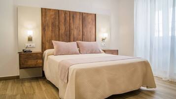 Un dormitorio con cabecera de madera, dos mesitas de noche con lámparas, una cama con colcha color beige y piso de madera.