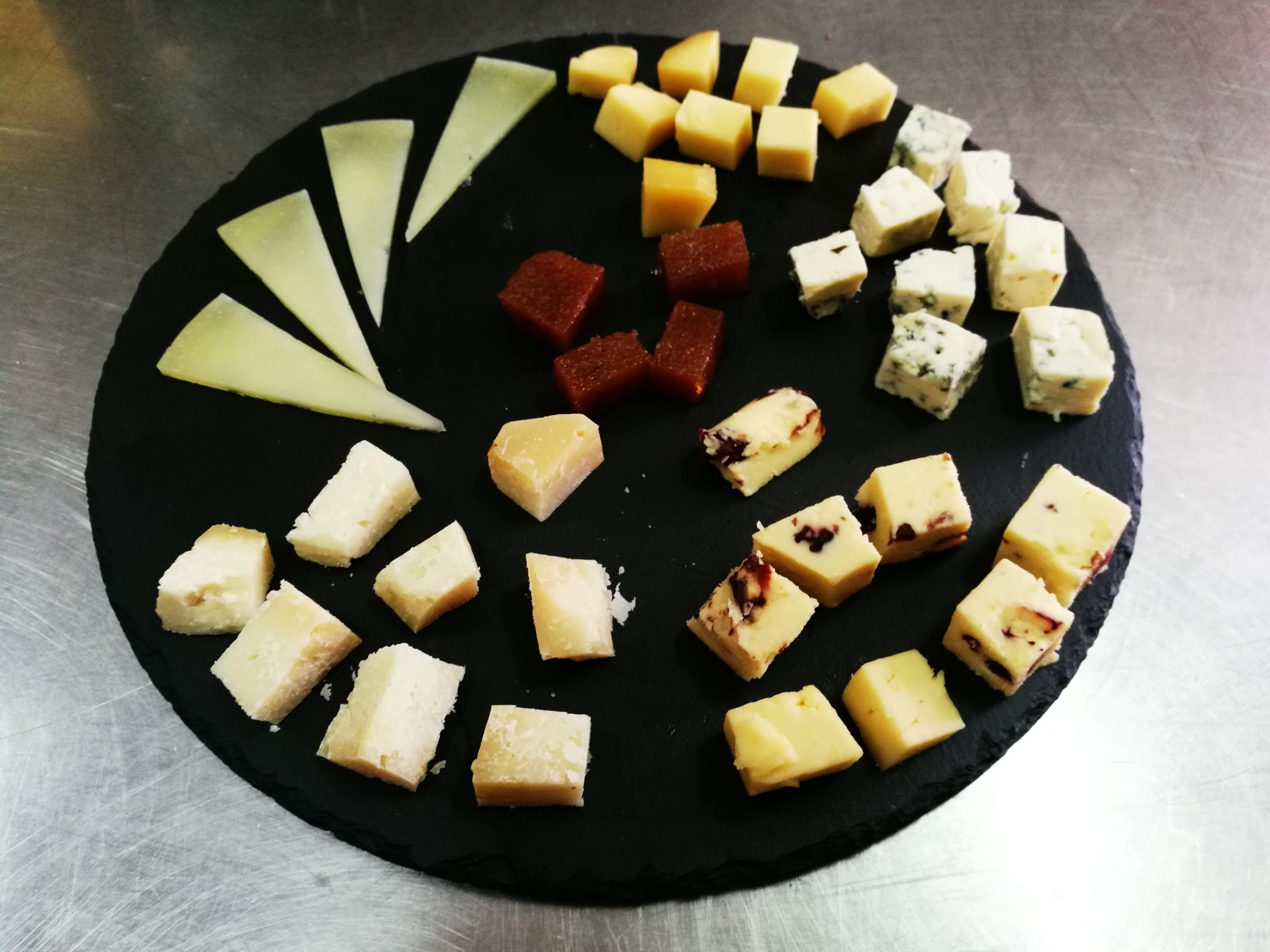 Una tabla redonda de pizarra negra con varios tipos de queso y fruta dispuesta sobre ella.