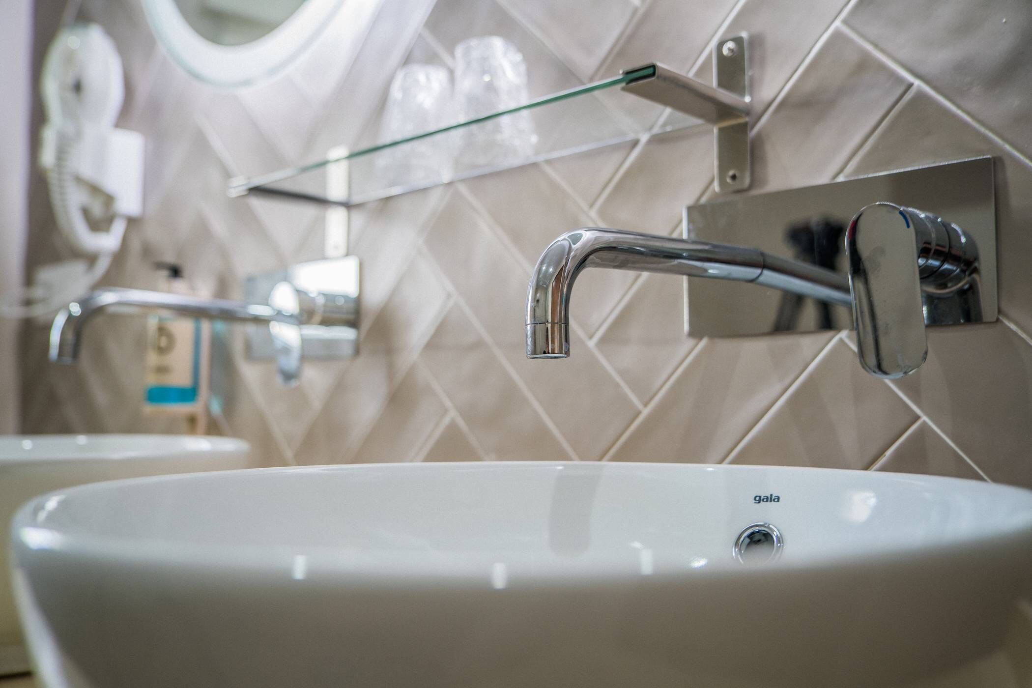 Un baño con lavamanos blanco, grifo cromado y estante de vidrio sobre el espejo.