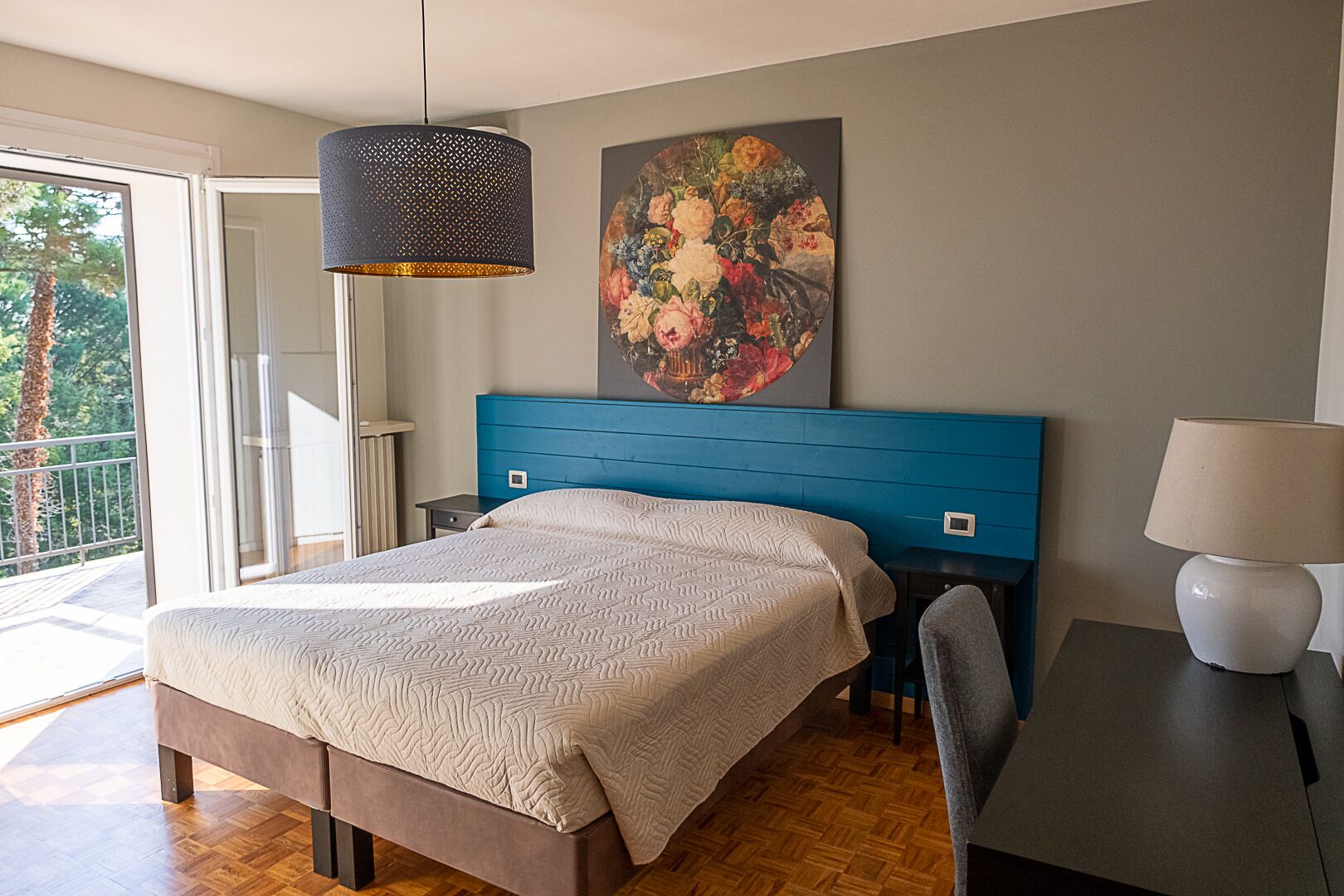 A bedroom with a large bed, a blue headboard, a hanging light fixture, a painting, and a view of the outdoors.