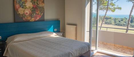A bedroom with a bed, a nightstand, a bench, a large painting, and a sliding glass door leading to a balcony with a view of trees and the sea.