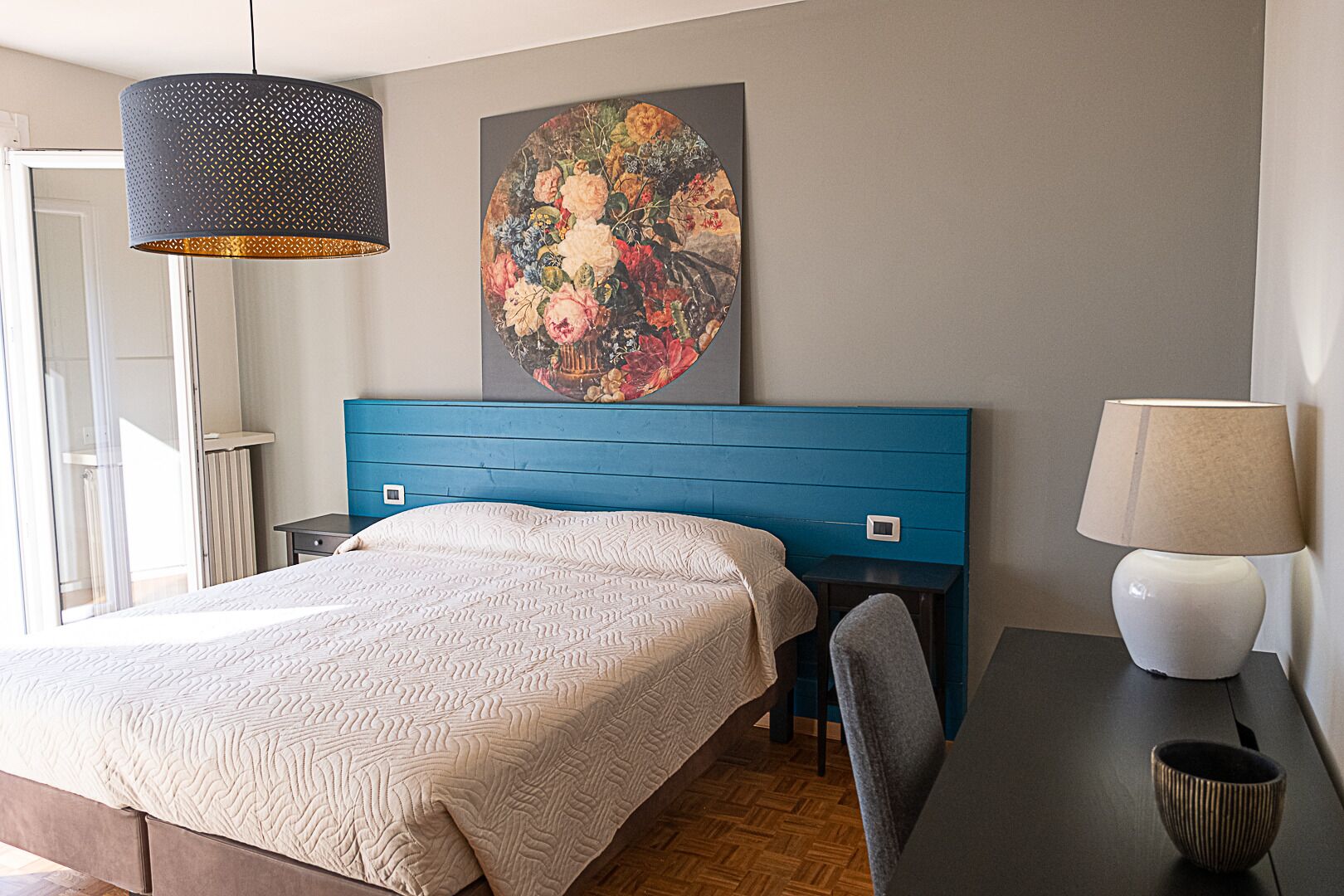A bedroom with a bed, a blue headboard, a painting, a lamp, and a chair.