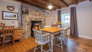 Une pièce chaleureuse avec une cheminée en pierre, des poutres apparentes en bois au plafond, une table à manger en bois entourée de chaises blanches et une fenêtre agrémentée de rideaux.