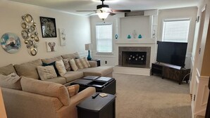 Un salon avec un canapé d’angle beige, une cheminée, un téléviseur et un ventilateur de plafond.