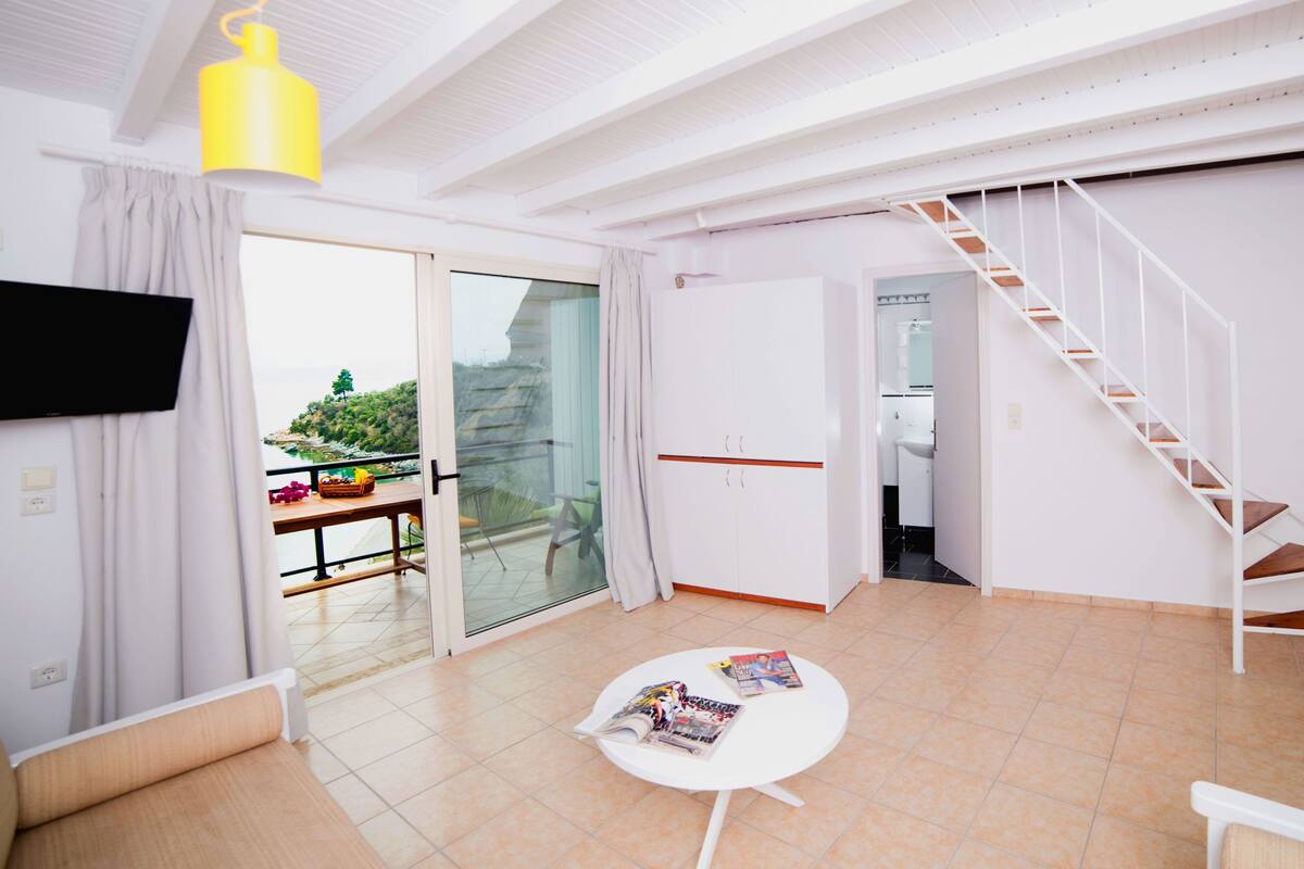 A modern living room with a flat-screen TV, a round table with magazines, a staircase, and a balcony with a view of greenery.