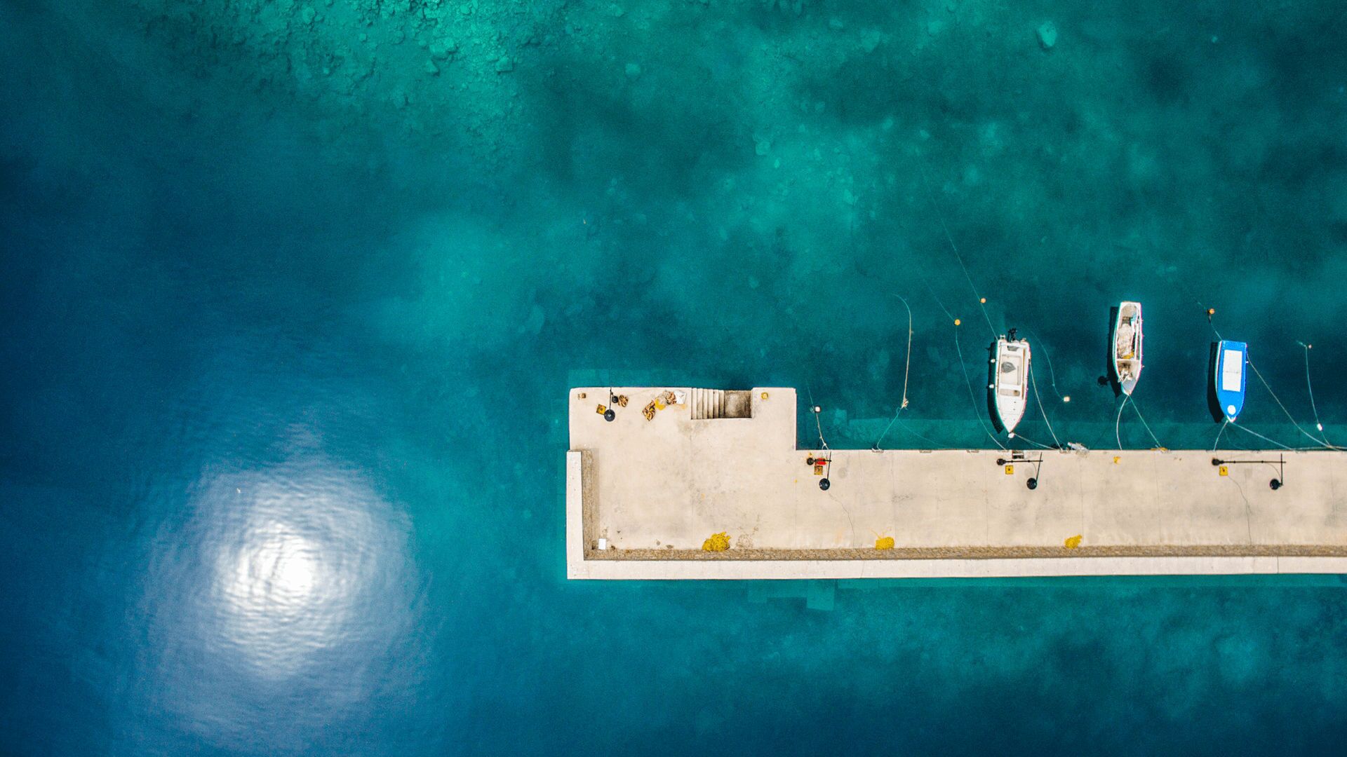 An aerial view of a pier with boats docked, clear turquoise water, and a concrete walkway.