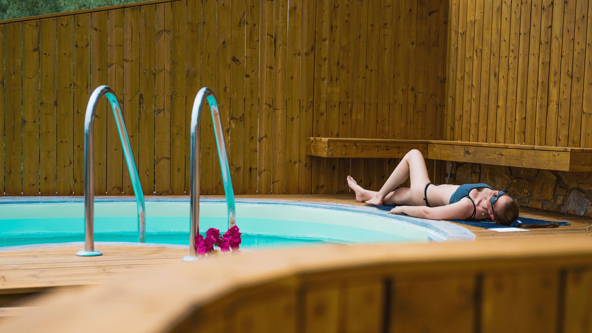 Une personne allongée sur une terrasse au bord d’une piscine, avec des rampes en bois et un mur de pierre en arrière-plan.