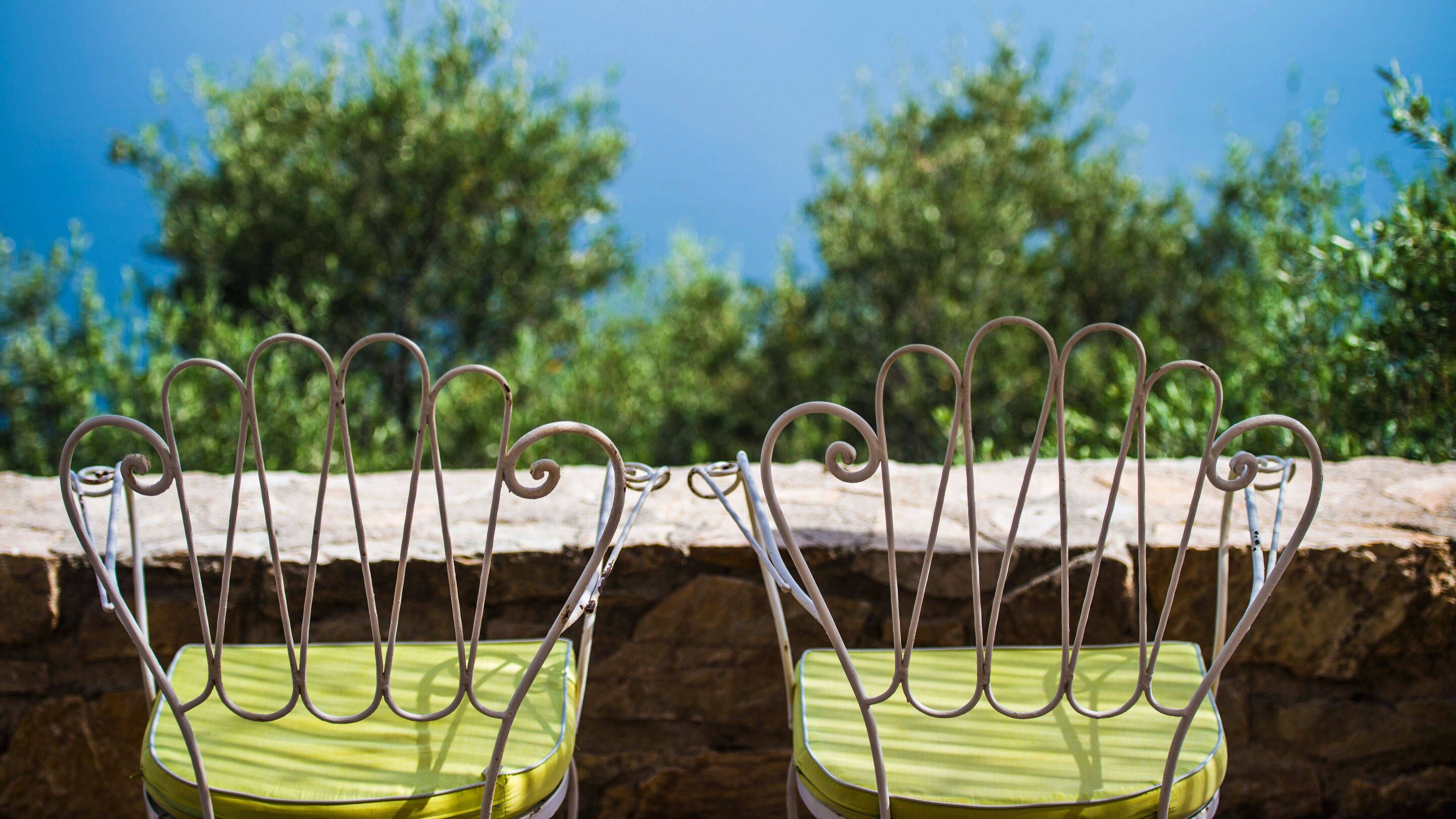 Two metal chairs with green cushions, positioned on a stone ledge with a stone wall and trees in the background.