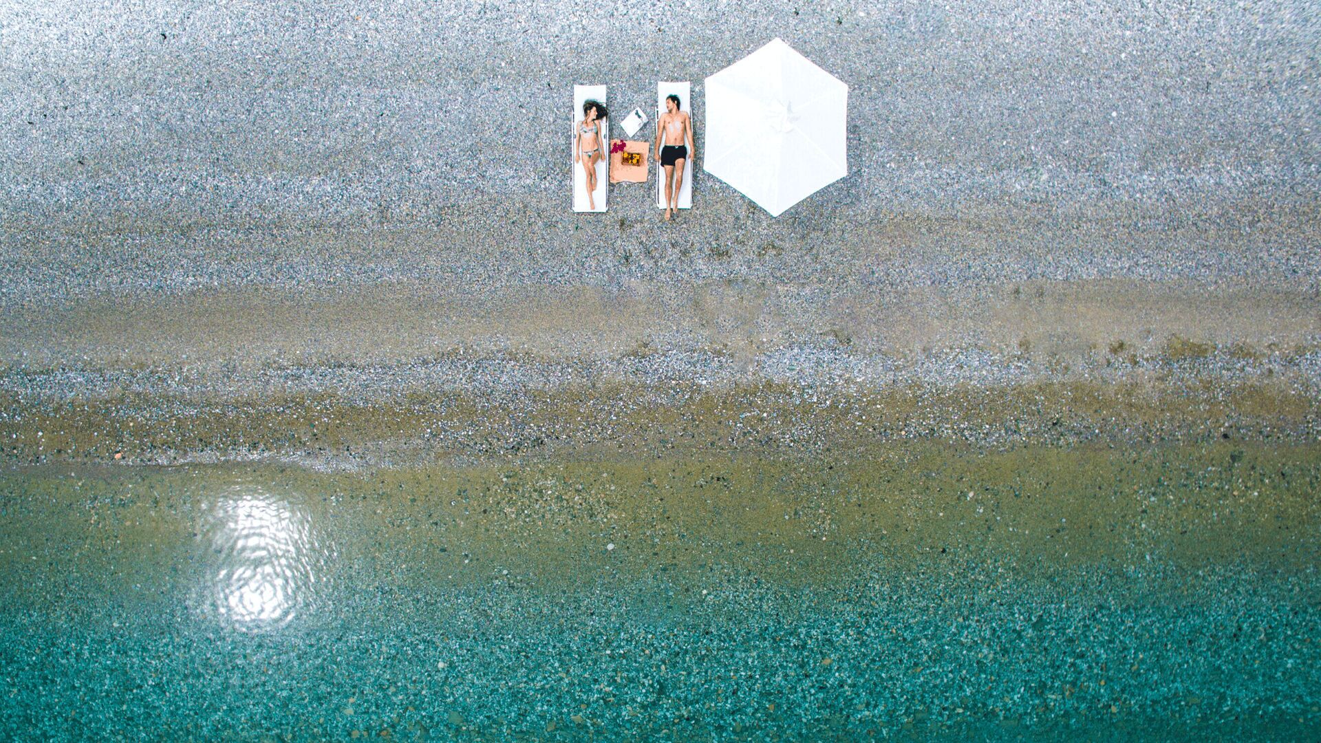An aerial view of a beach with two people lying on towels, a white hexagonal object, and a clear water body.