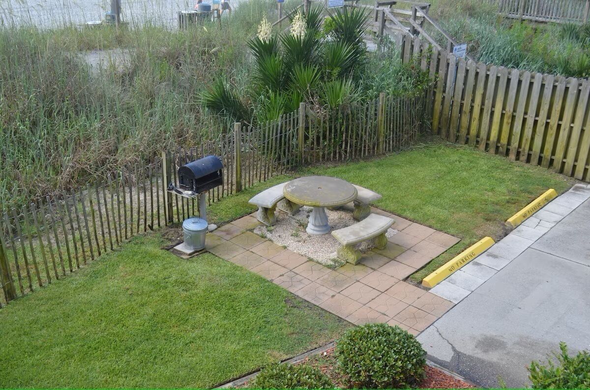 Un espace jardin agrémenté d’une table en pierre et de bancs, entouré d’une clôture en bois et d’une végétation luxuriante.